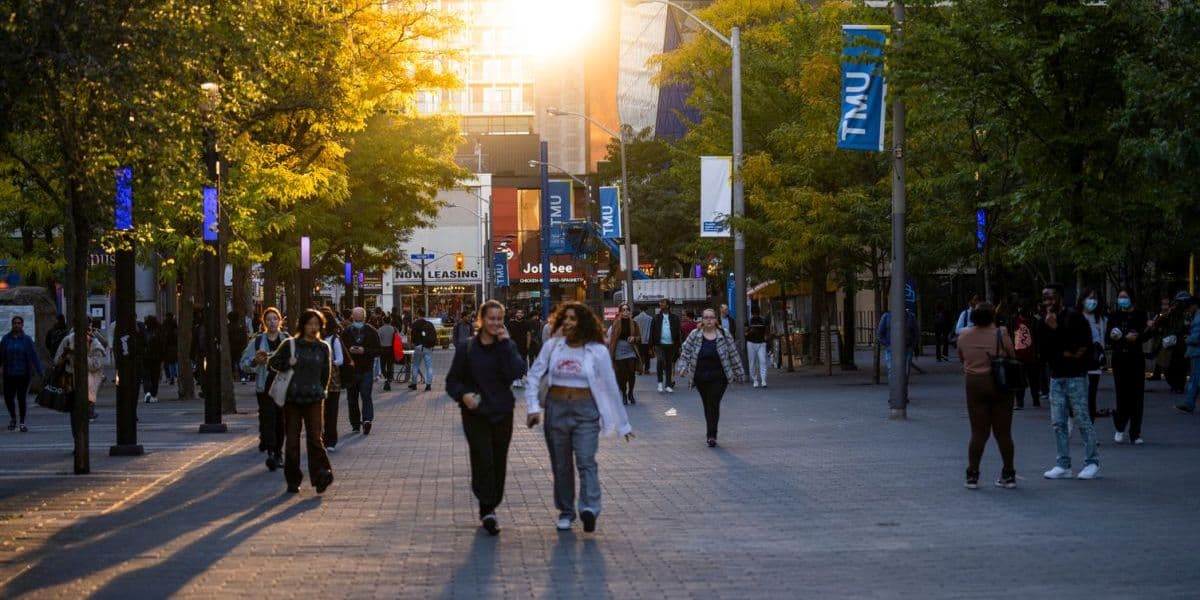 A street scene looking west on Gould Street towards Yonge Street. Many students are walking purposefully through the area. TMU banners are hanging from the streetlights and the setting sun is reflected in a building the distance.