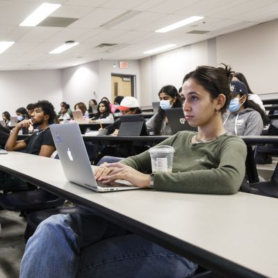 A white woman with dark hair sitting in a lecture hall, taking notes on her laptop.