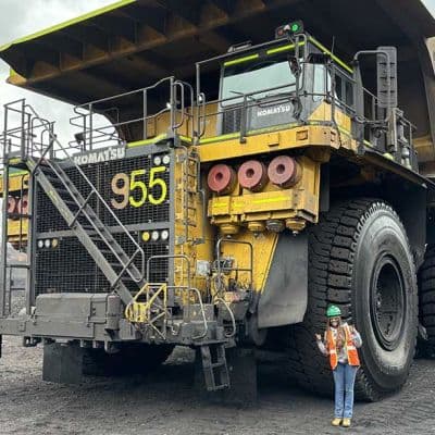 A Black woman wearing a hard hat and safety vest posing in front of a massive heavy machinery truck. The wheel of the truck is twice her height.