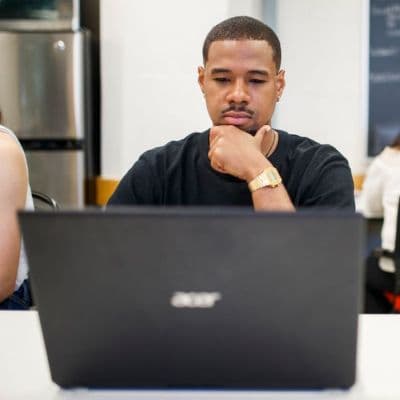 A Black man, staring intently at his laptop screen. He is resting his chin in his hand.