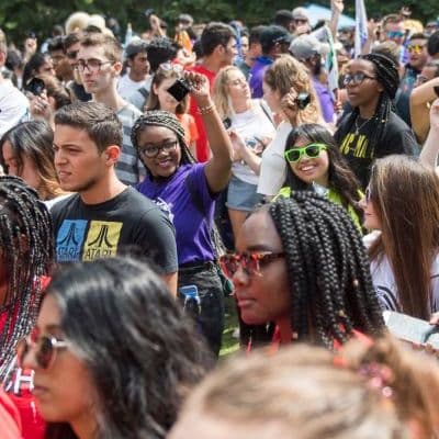 A crowd shot of an outdoor orientation event. A Black woman looks toward the camera through the crowd; she has her arm in the air. A woman beside her is wearing lime green sunglasses and smiling.