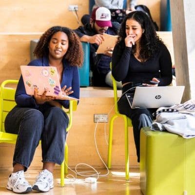 A Black woman and a white woman sitting beside each other in the Student Learning Centre. They both have laptops open, and the Black woman is showing something on her screen. The white woman is laughing with her hand in front of her mouth.
