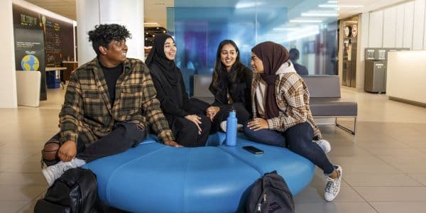 Four students - a man and three women - are sitting on a circular padded bench in the Service Hub waiting area. They are facing each other, talking and smiling. Two of the women are wearing hijabs.