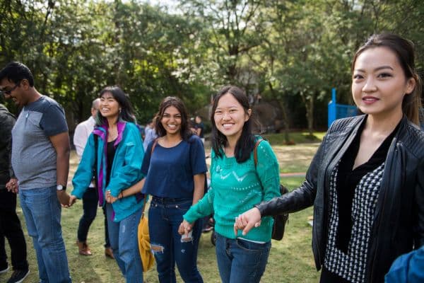 A group of young Asian women, engaged in an activity in the outdoor Quad. They are all in a line, holding hands and laughing.