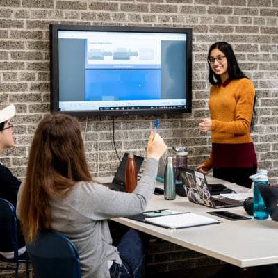 A meeting room, where one young woman is standing up in front of a large screen with a presentation on it. A man and a woman are sitting at a table with several laptops on it, asking the woman questions.