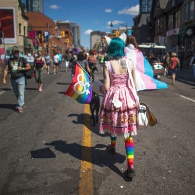 A person is walking away from the camera on a city street. They have bright blue hair, and are wearing cat ears, a pink dress, and rainbow knee socks. They are carrying a lesbian pride flag.