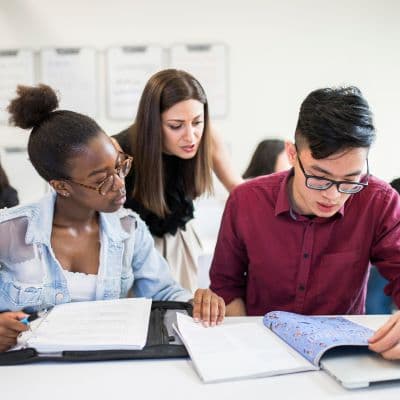 An Asian man is looking at a notebook. A Black woman sits beside him, also looking at the notebook. A white woman is standing behind them, talking to them. 
