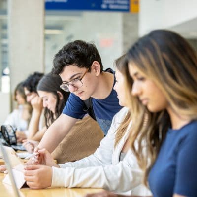 A side view of students working at a long desk. The focus is on a white woman, who is showing something on her laptop to a white man with glasses, who is leaning over her shoulder. 