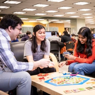 A white man, a white woman and a South Asian woman are sitting around a board game. They are holding cards and appear to be having fun playing.