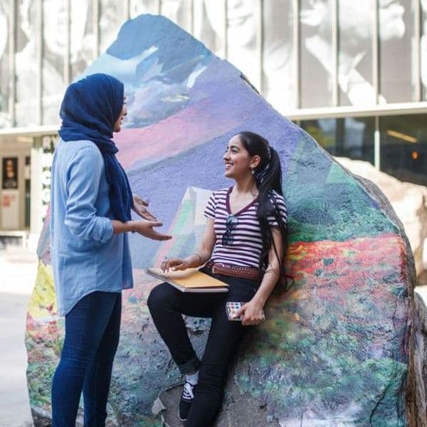 Two women are talking, one is leaning on a brightly painted rock in an empty Lake Devo. One woman is wearing a hijab.