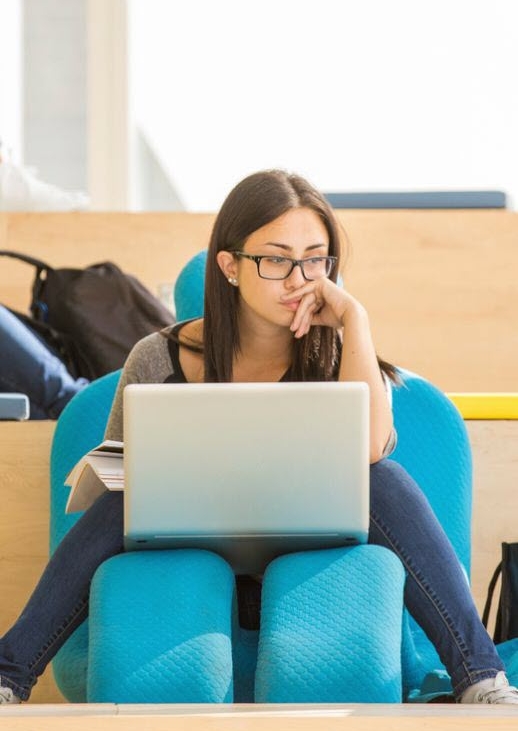 A woman sitting on a lounge chair in the Student Learning Centre. An open laptop and a textbook are balanced in front of her, but she is looking into the distance, as if she is thinking. 
