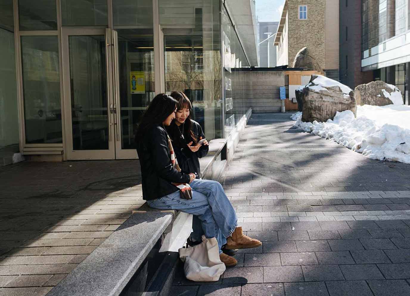 Two students sitting on a bench on TMU campus in the winter, checking phone for loan status