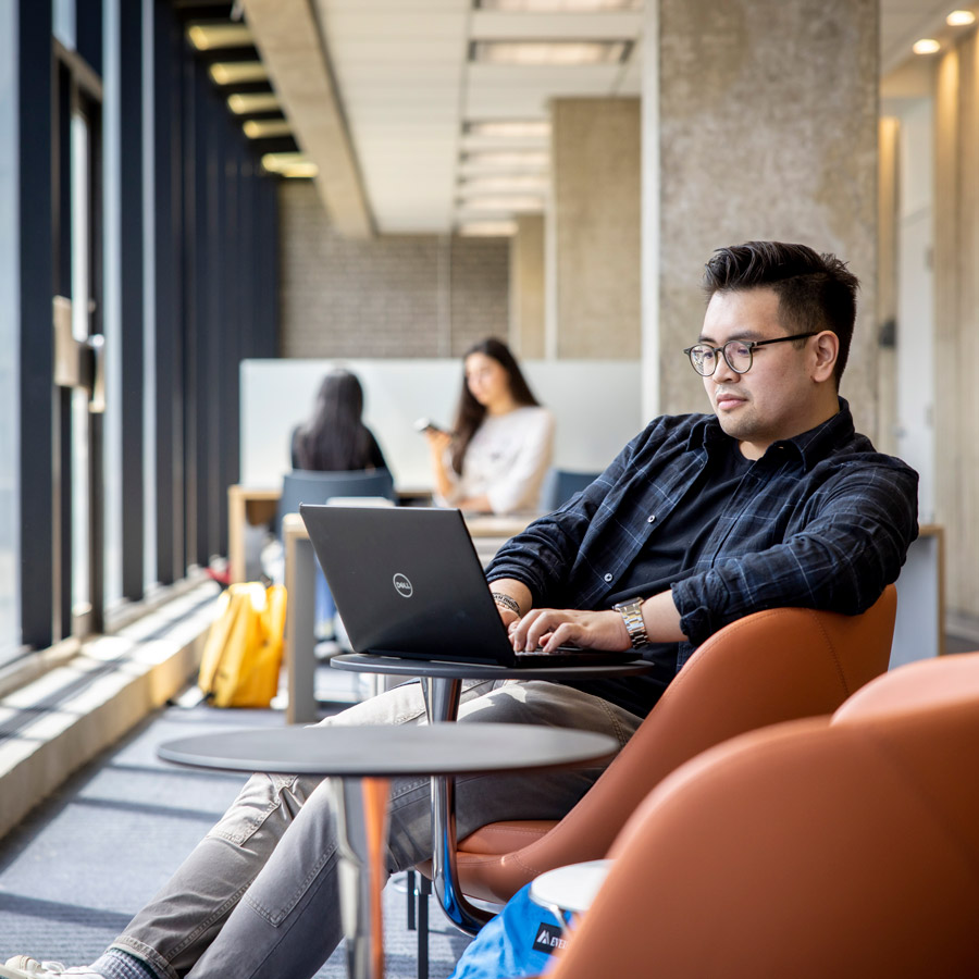 A student casually leans back in an armchair in the SLC while working on a laptop.