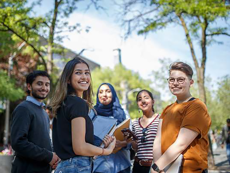 Diverse group of five students smiling on campus. One of them is wearing a hijab.