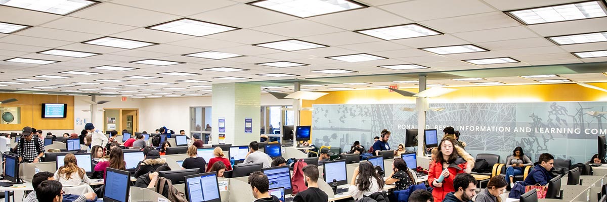 The square ceiling light panels in one of the Library common areas with many students working below on computer workstations.