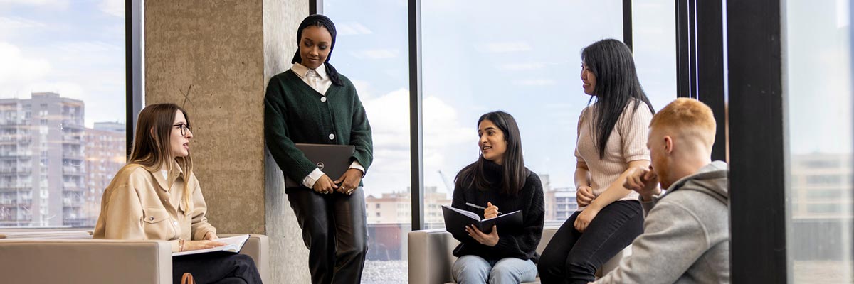 A group of TMU students sit chatting in a room with lots of windows showing a view of downtown Toronto.