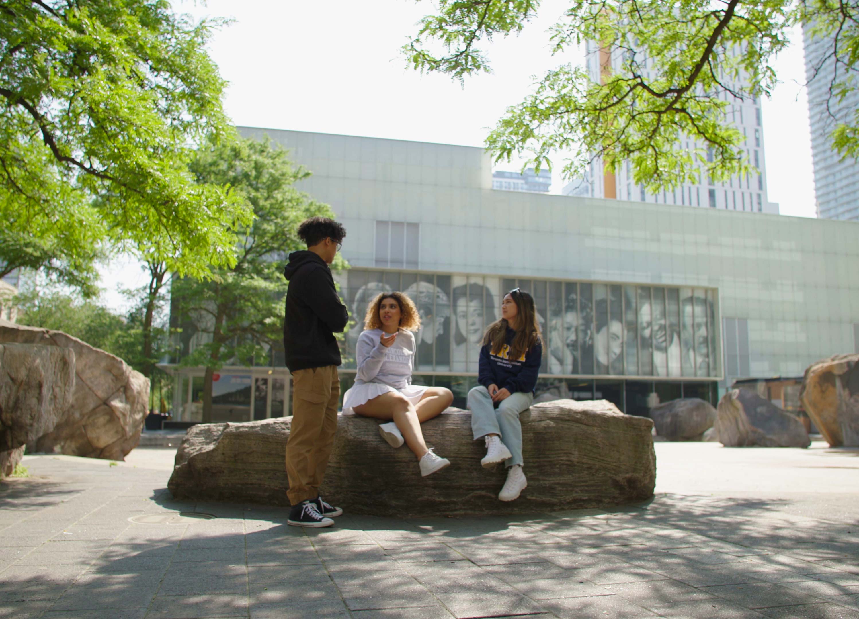 Three students chatting beside Lake Devo, with two of them sitting on the rocks.
