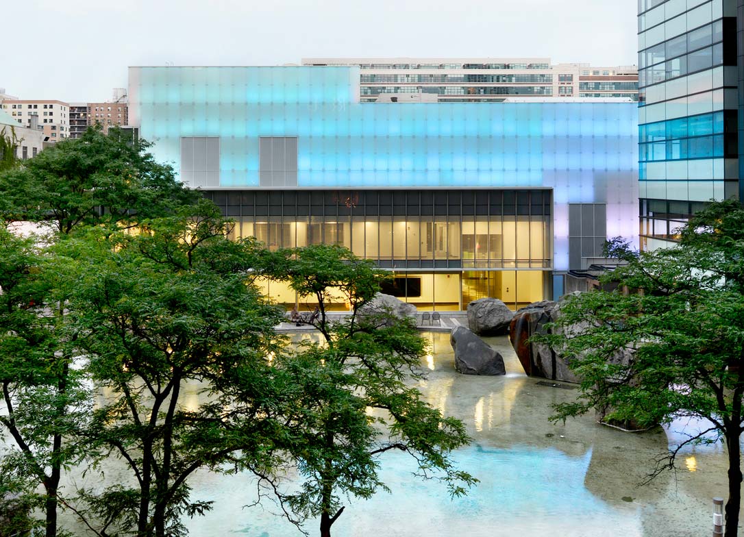 The Image Centre lit up with light blue lights, reflecting onto Lake Devo flanked with green trees.
