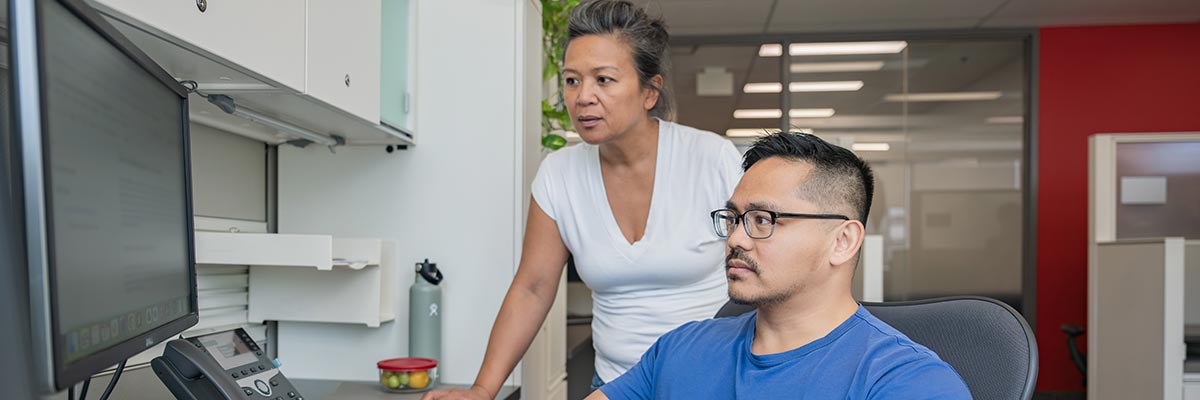 Two employees collaborate at a computer, one of them sitting and the other looking over his shoulder.