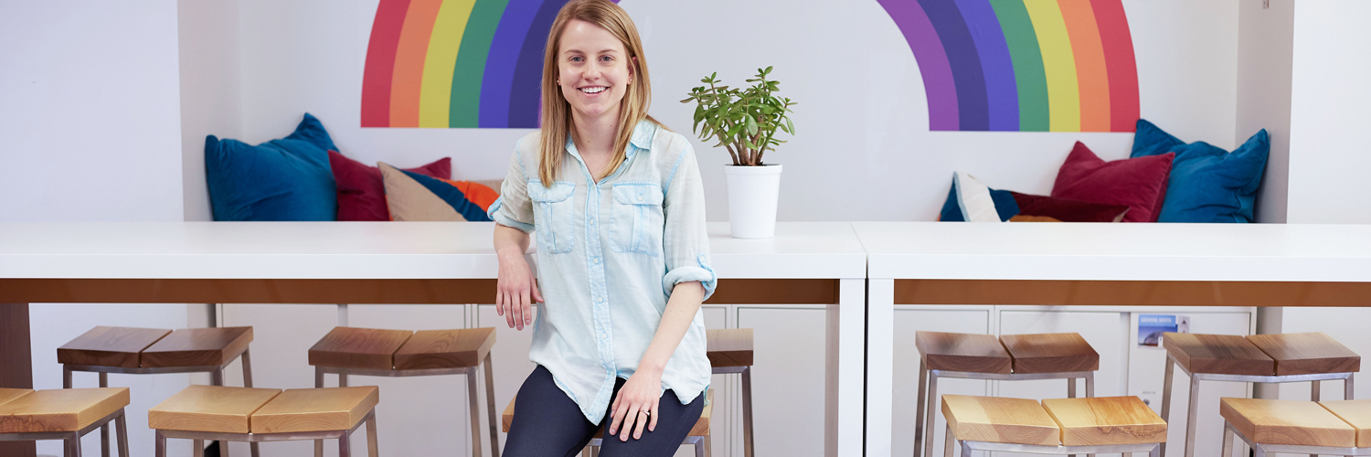 Entrepreneur smiling directly at camera and sitting on a stool, with elbow resting on a table 