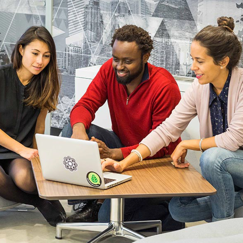 entrepreneurs gathered around table, looking at computer and smiling 