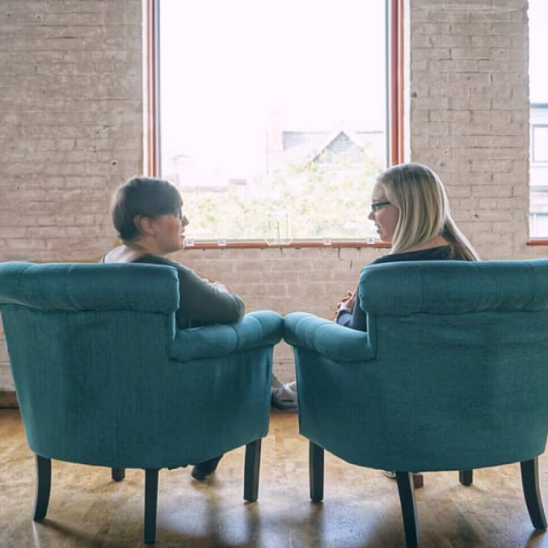 Anne and a woman sitting in blue chairs and speaking, looking away from the camera