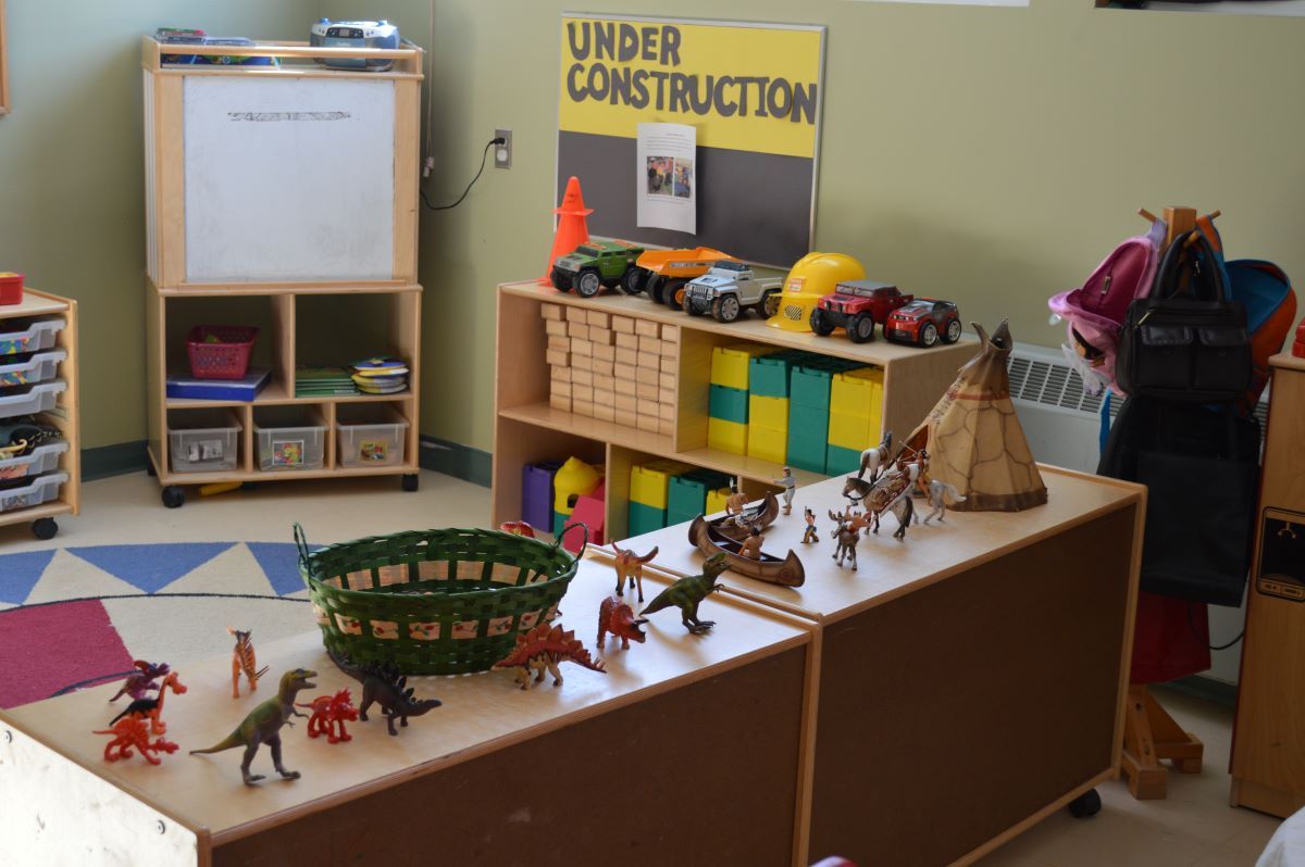 Une salle déserte dans un centre de garde d’enfants. Des tablettes en bois remplies de jouets pour enfants et une affiche jaune et noire sur laquelle est inscrite « Under Construction ». Un tableau blanc, une aire de cercle et un portemanteau avec des sacs à dos. 