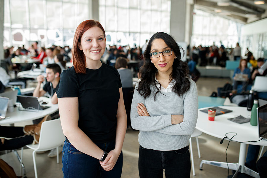 Two students are standing on the 8th floor of SLC on a busy day. They are looking at the camera. Other students can be seen working in the background. 