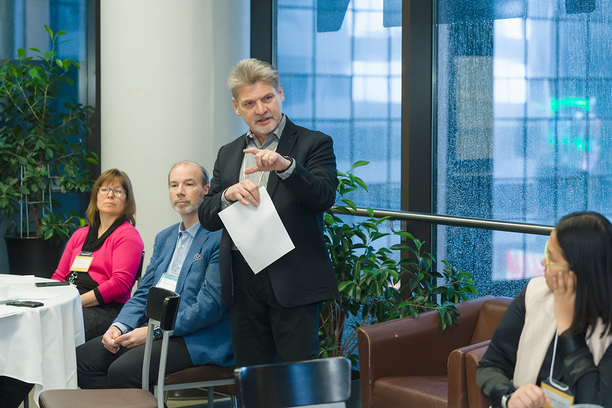 A man stands gesturing with a paper in hand, addressing seated attendees at the advisory council meeting event. Attentive expressions, indoor setting with large windows and plants.