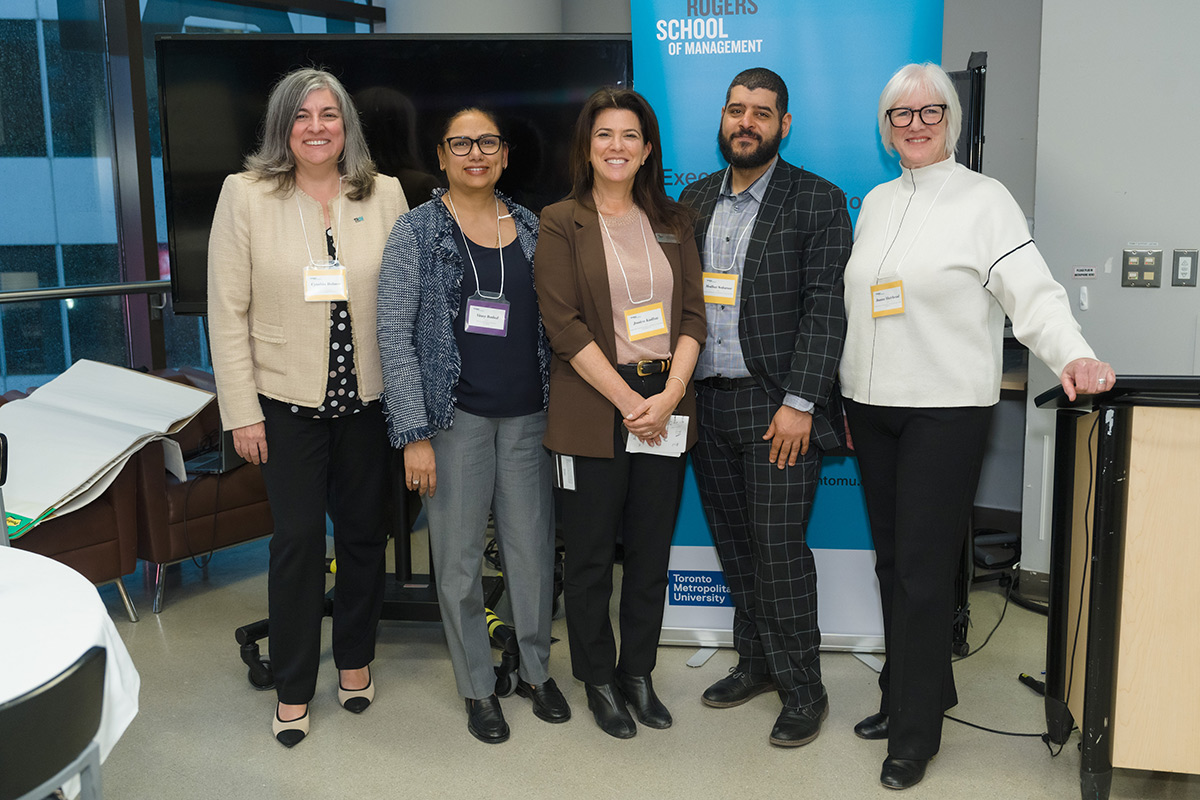 Dean Cynthia Holmes with staff and advisory council members stand smiling in front of a blue Toronto Metropolitan University banner at the advisory council meeting event.