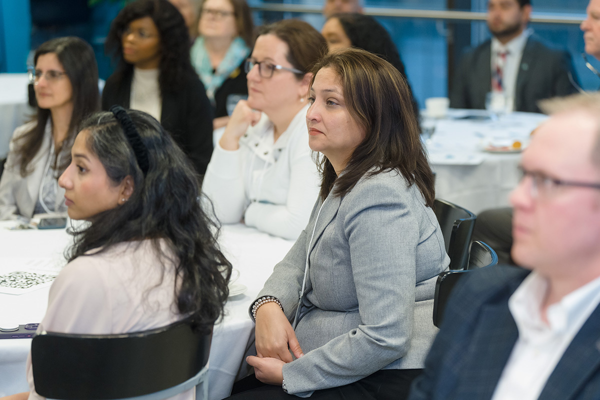 A diverse group of people attentively listens at the advisory council meeting event. The setting is formal, with participants seated at round tables.
