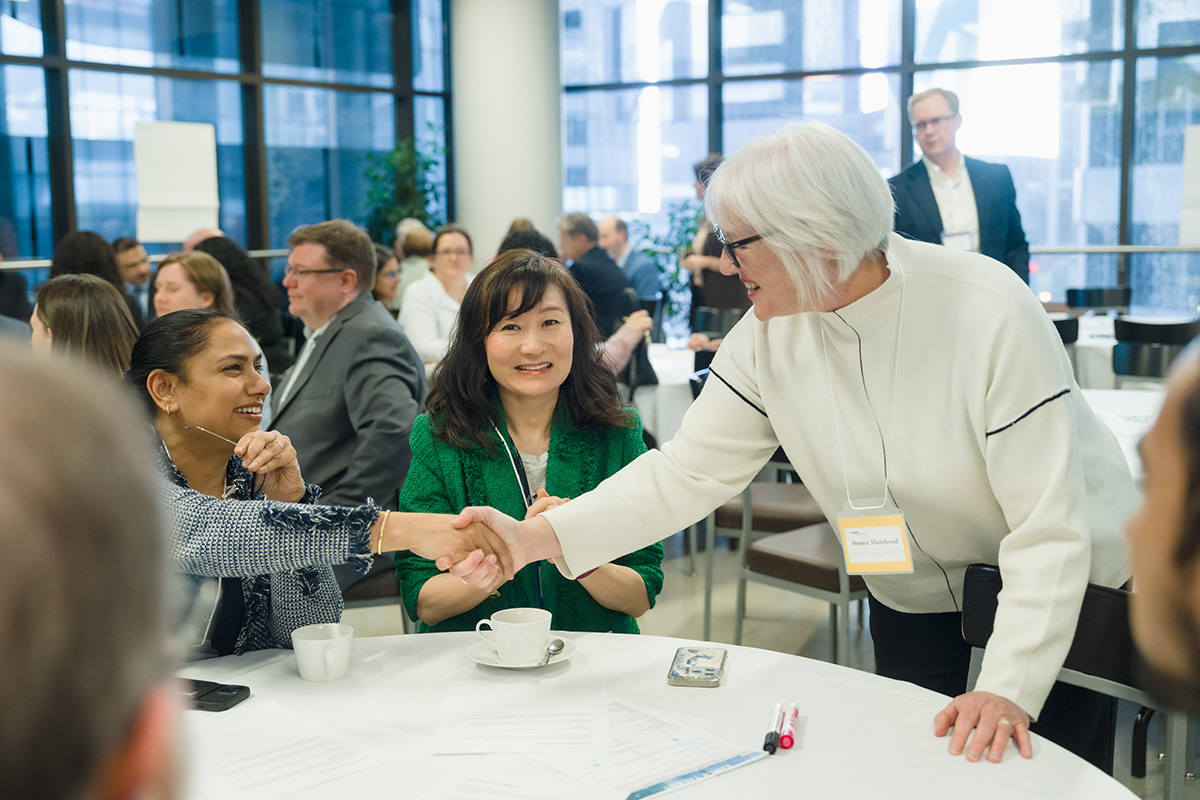 A diverse group at a conference table engages warmly. Two women in focus shake hands, smiling. The atmosphere is friendly and professional.