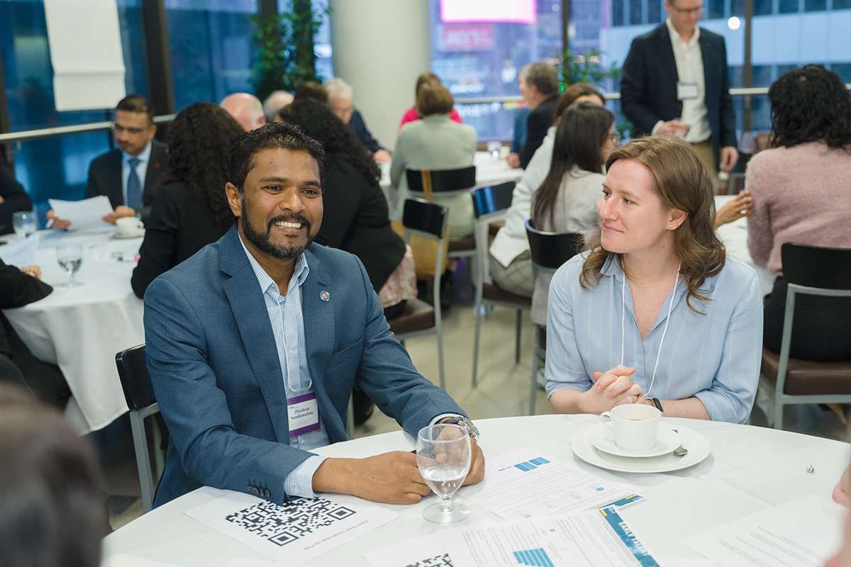 A diverse group of professionals engages in discussion around tables at the advisory council meeting. A smiling man in a blue blazer and a woman with a cup lead the conversation.