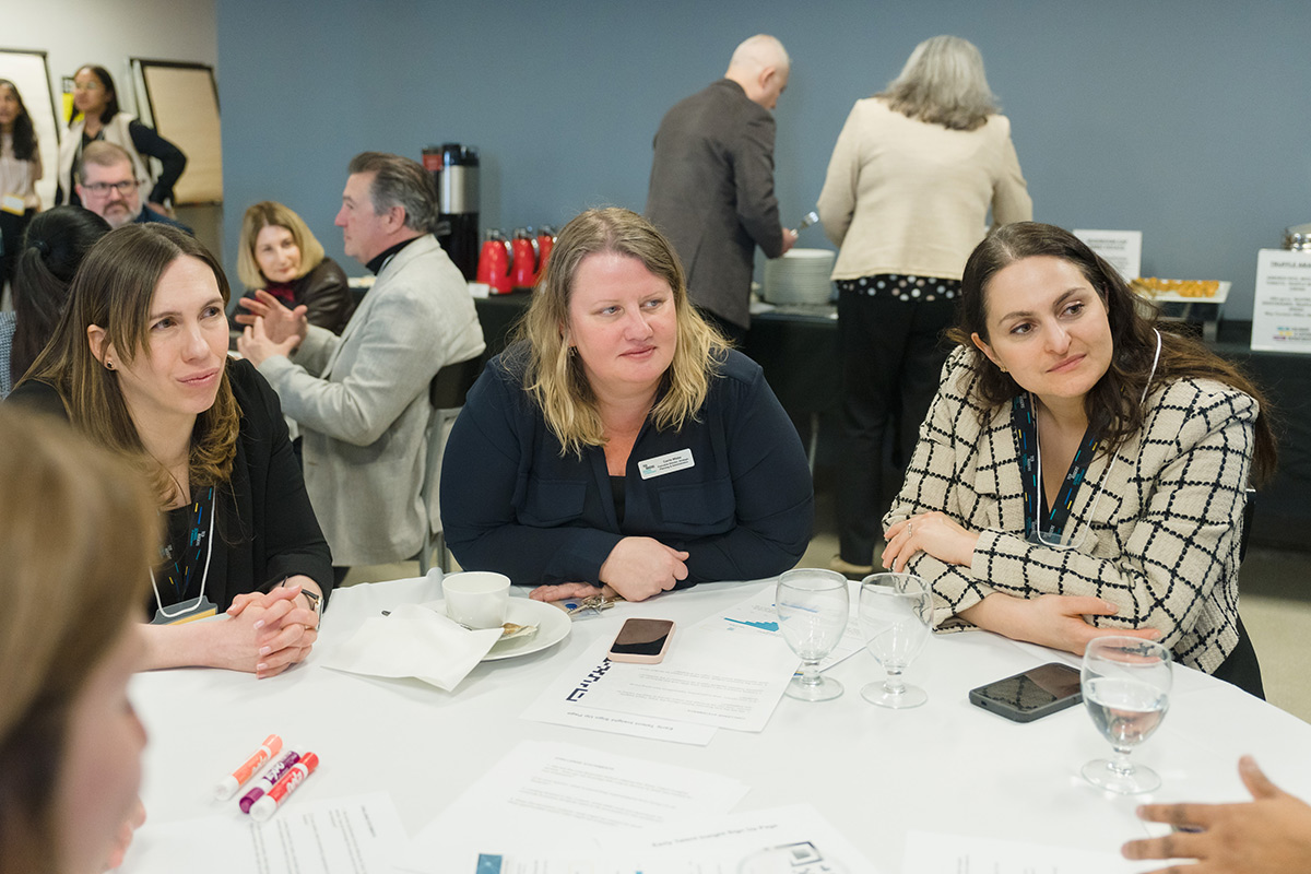 Three women engaged in conversation at a round table during a meeting. Papers and markers are on the table. The setting is relaxed and professional.