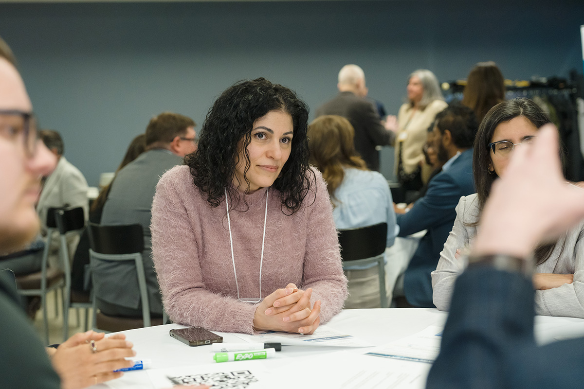 Rita Loncar sits attentively at a round table during a discussion. People converse in the background. Professional and engaged atmosphere.