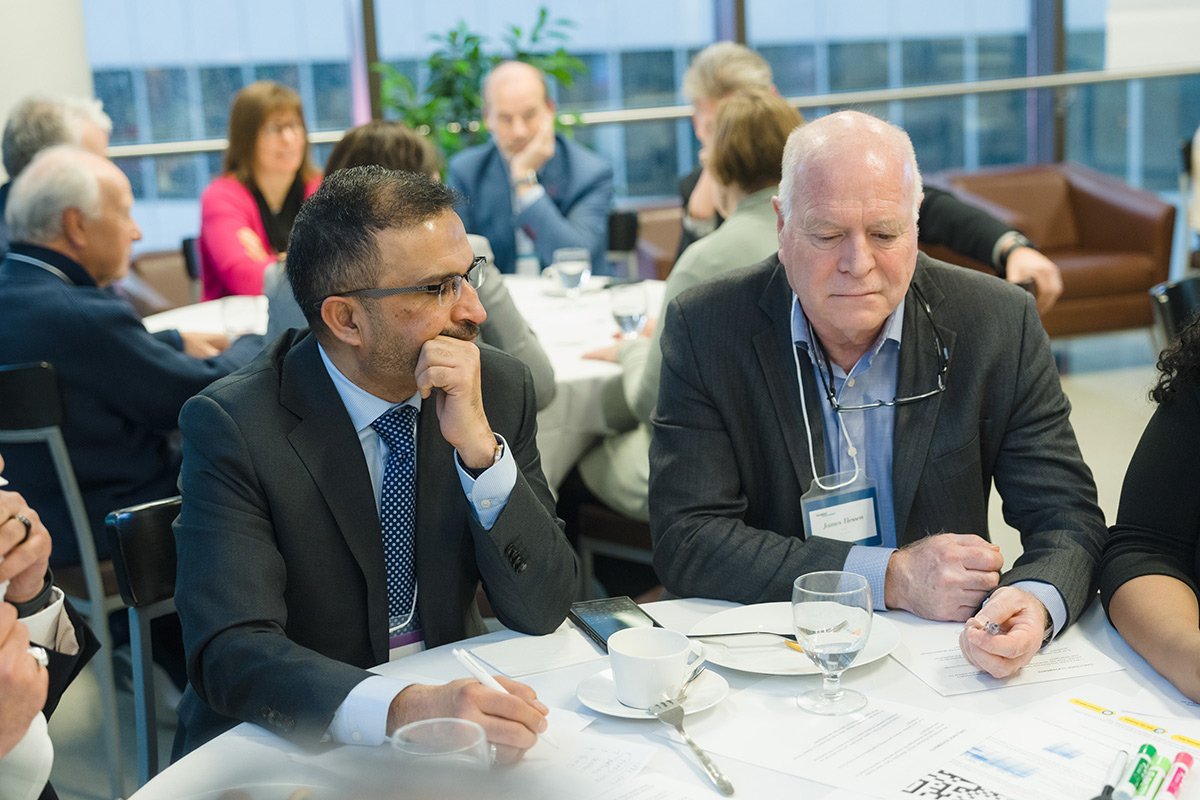 A group of professionals in business attire are engaged in discussion around a table at the advisory council meeting. Papers, cups, and glasses are on the table, indicating a formal meeting. The mood is serious and focused.