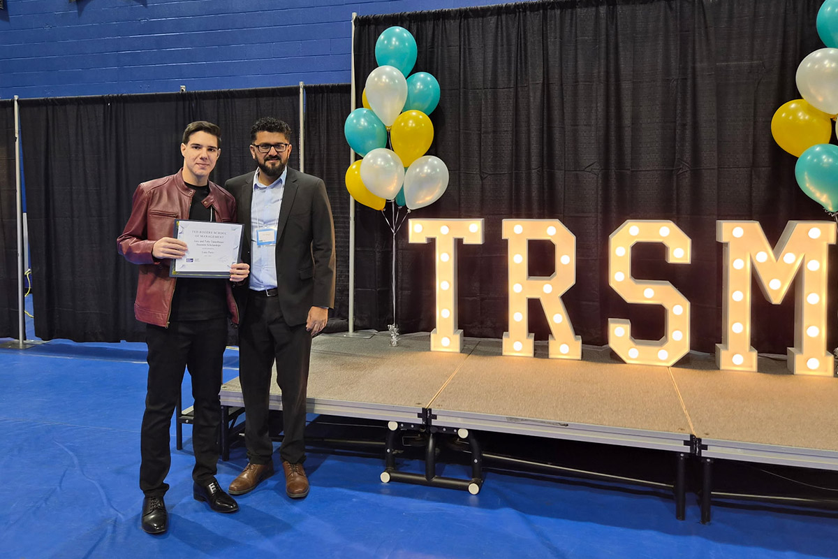 Professor Vik Singh and a student stand on a stage against a black curtain backdrop. The student holds a certificate. Large lit-up letters spell "TRSM" beside colourful balloons.