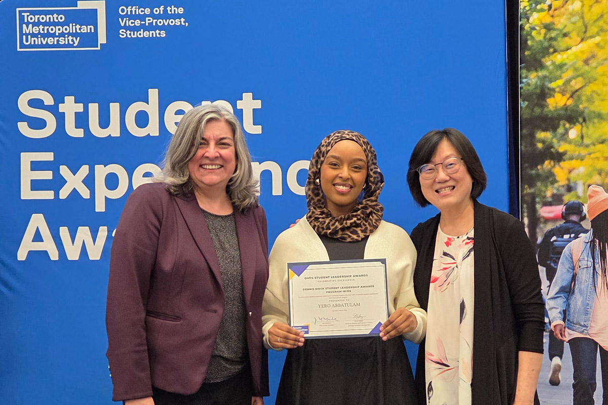 A student holds a certificate, while standing in the middle of Dean Cynthia Holmes and professor Clare Chua-Chow