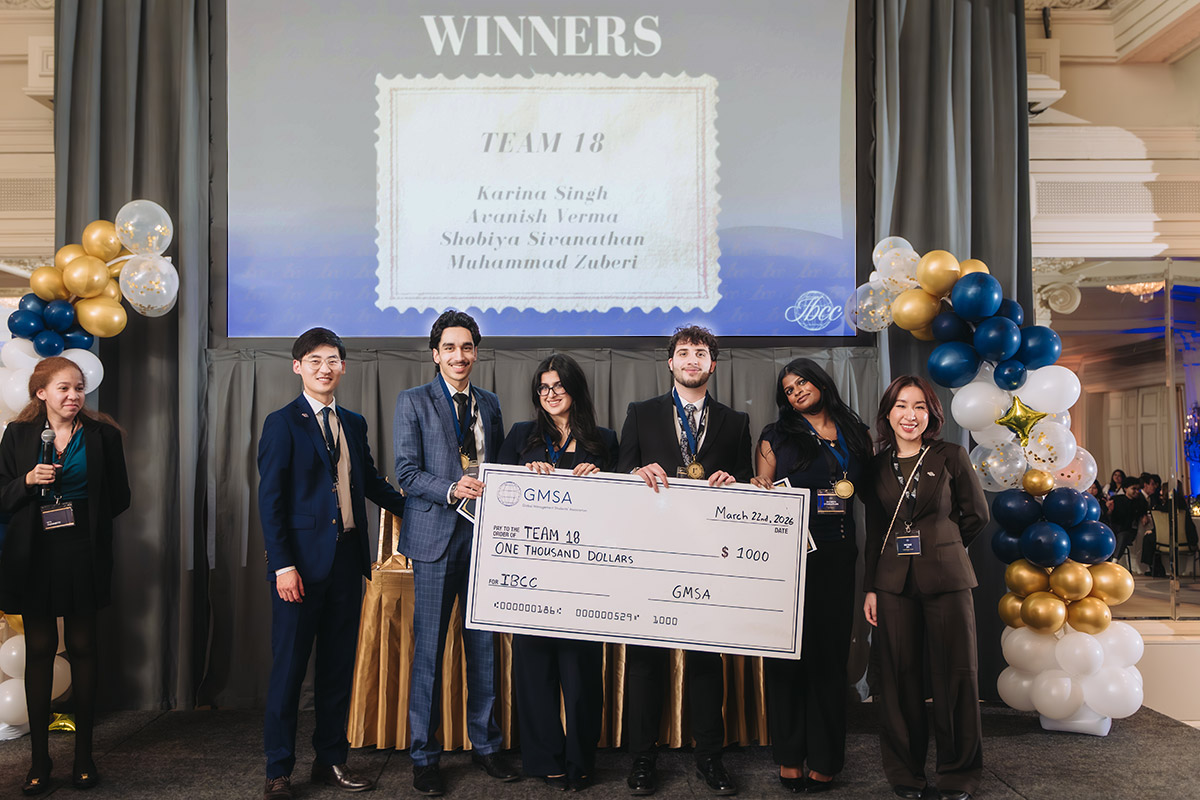 GMS students stand onstage, smiling and holding an oversized check for 1,000 dollars. A display overhead shows "Winners, Team 18." Balloons frame the stage.