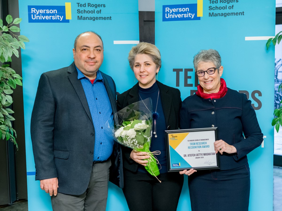 One man and two women stand in front of school banner holding award