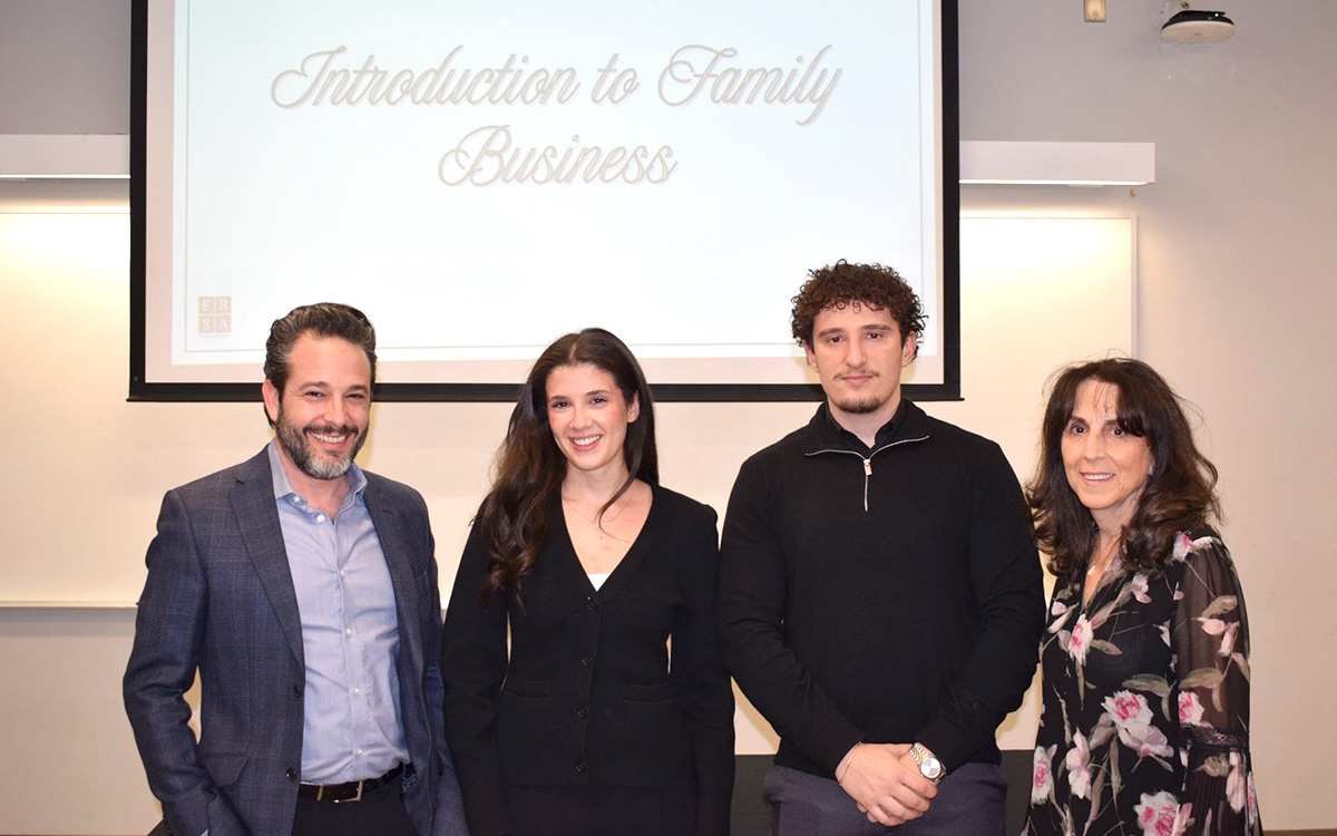 Professor Francesco Barbera, two FBSA students, and Janie Goldstein stand smiling in a classroom setting under a projection reading "Introduction to Family Business." They appear professional and engaged.