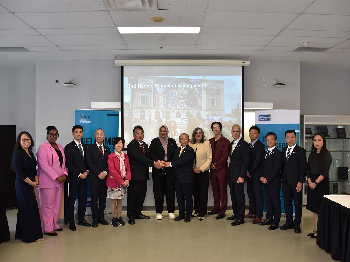 During a Japanese delegation visit, 16 attendees stand for a photo in a row in front of a projector screen with "Ted Rogers School of Management" displayed in white block text behind them on a projector screen
