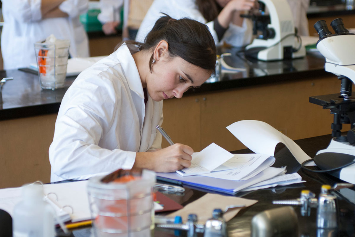 Student in a white lab coat writing notes for labwork.