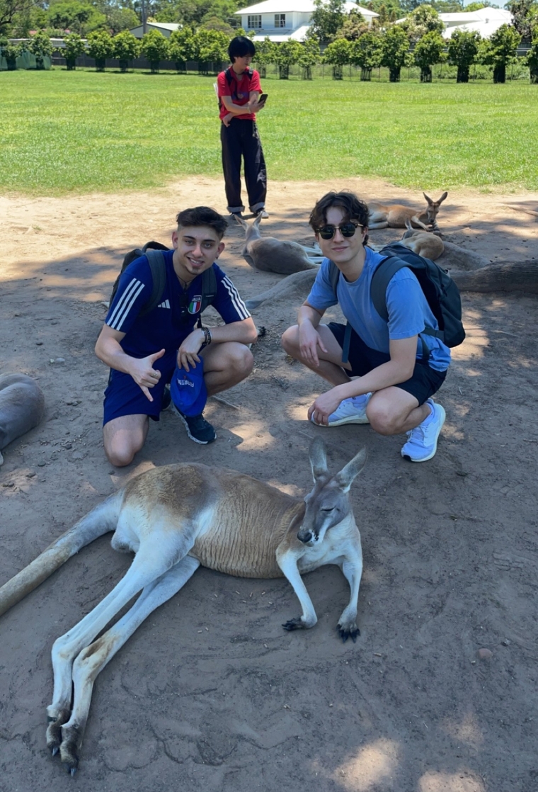 two students taking a picture with a kangaroo on the ground outside