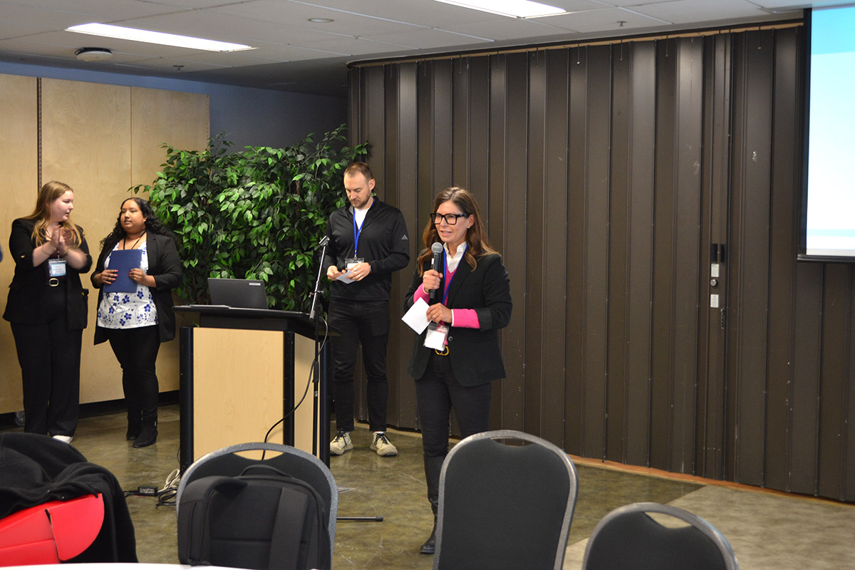 A woman in a suit speaks into a microphone at the CUSC conference, holding notes. Three colleagues stand by, two applauding.