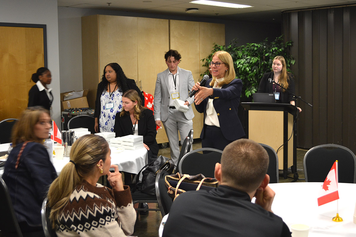 Karen Peesker in business attire speaks enthusiastically into a microphone at the SCUC conference. Attendees seated at round tables with Canadian flags, listening attentively.