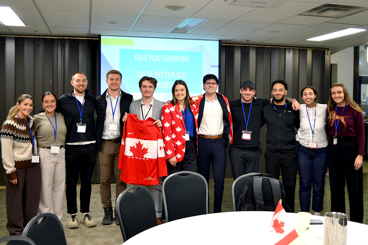 A group of 11 individuals smiling, pose for a photo. One person holds a red Canadian flag and two other are weating Canadian flag jackets. A screen in the background reads “Role Play Champion.” Celebratory mood.