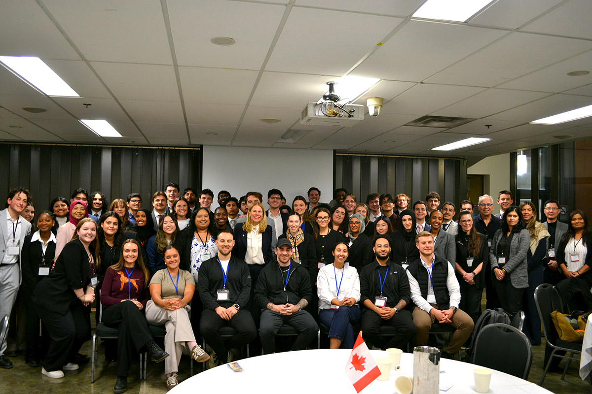 A large and diverse group of students smile for a group photo at the Canadian University Sales Competition (CUSC), with a small Canadian flag on a table in front of them.
