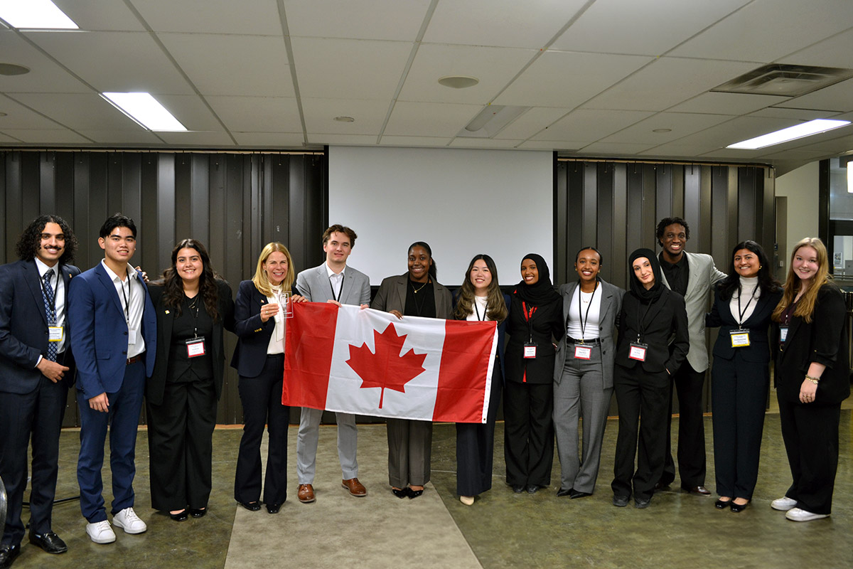 Karen Peesker with 12 students in formal attire stand smiling, holding a Canadian flag at TRSM. The atmosphere is celebratory and united.