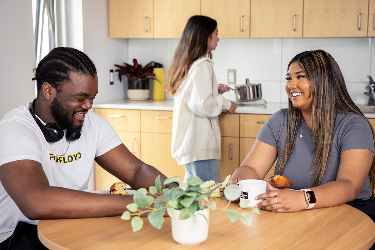 Two students at a table eating and talking, while another is walking behind them with a cooking pot